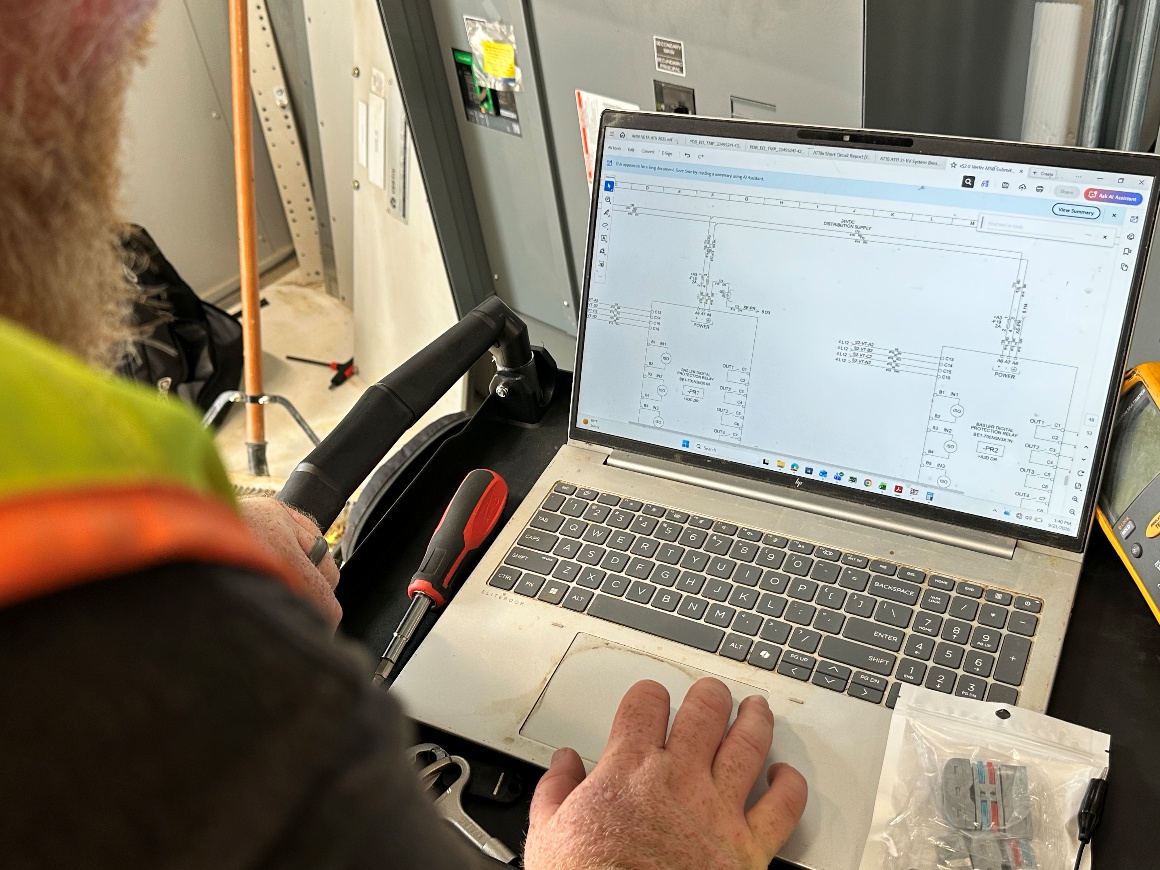 A technician compares electrical drawings on a laptop with physical device connections.